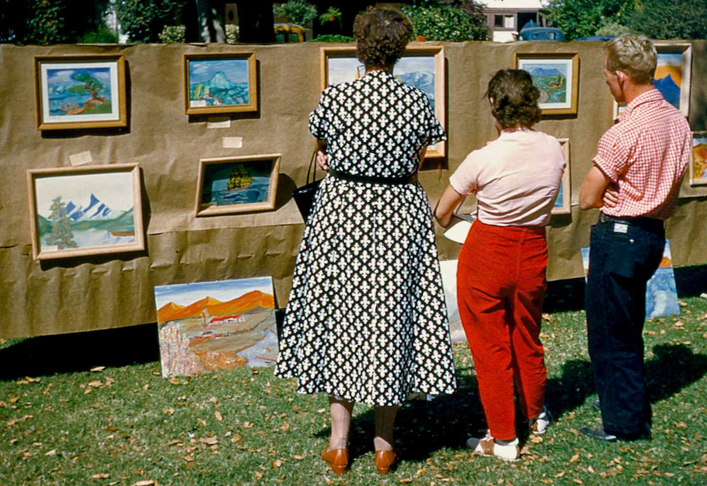 A view from behind three persons (2 females and one male) in contrapposto poses viewing an outdoor neighborhood art exhibit. Cira 1950's.